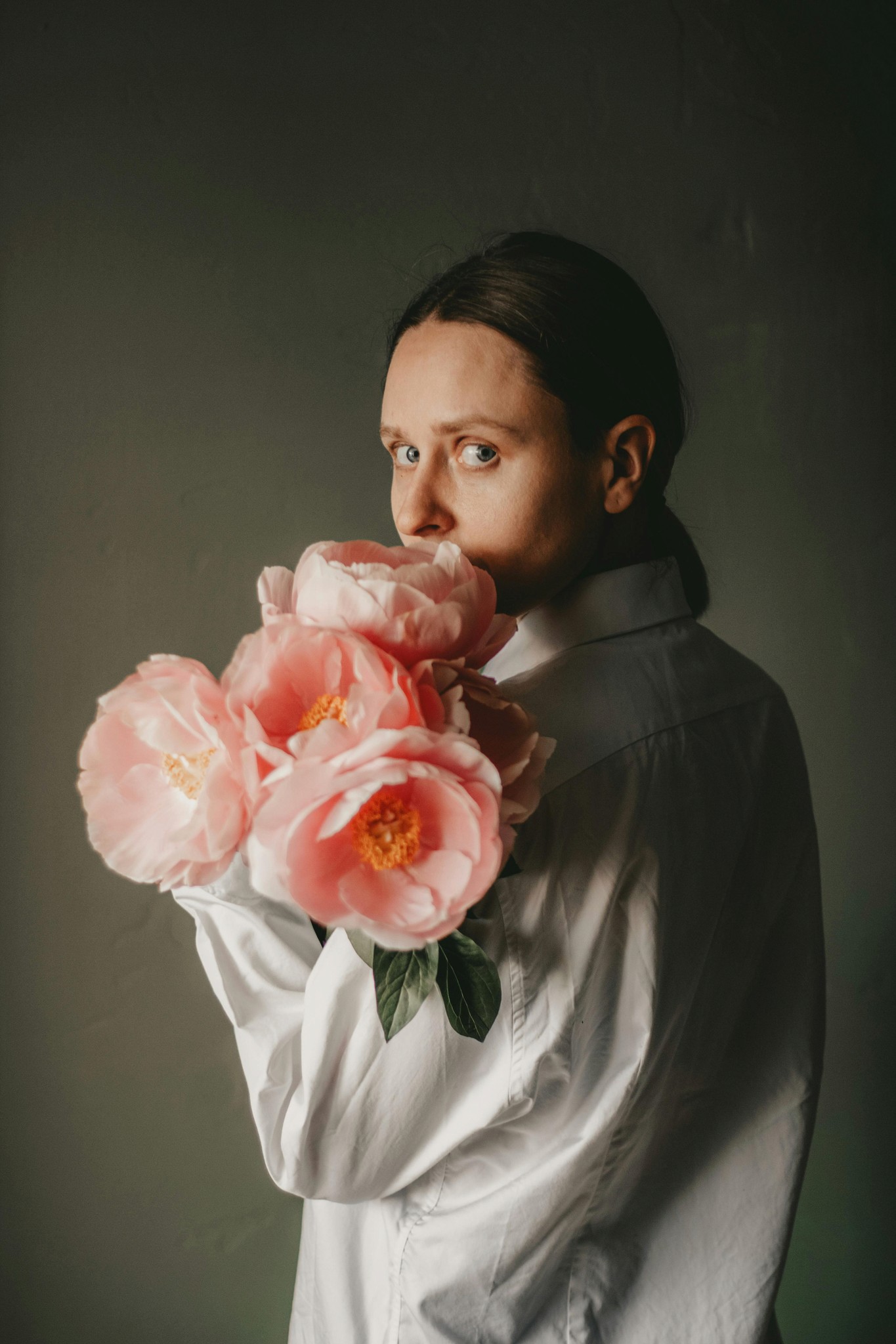 A person in a white shirt peering over a bouquet of large pink peonies against a dark green background.