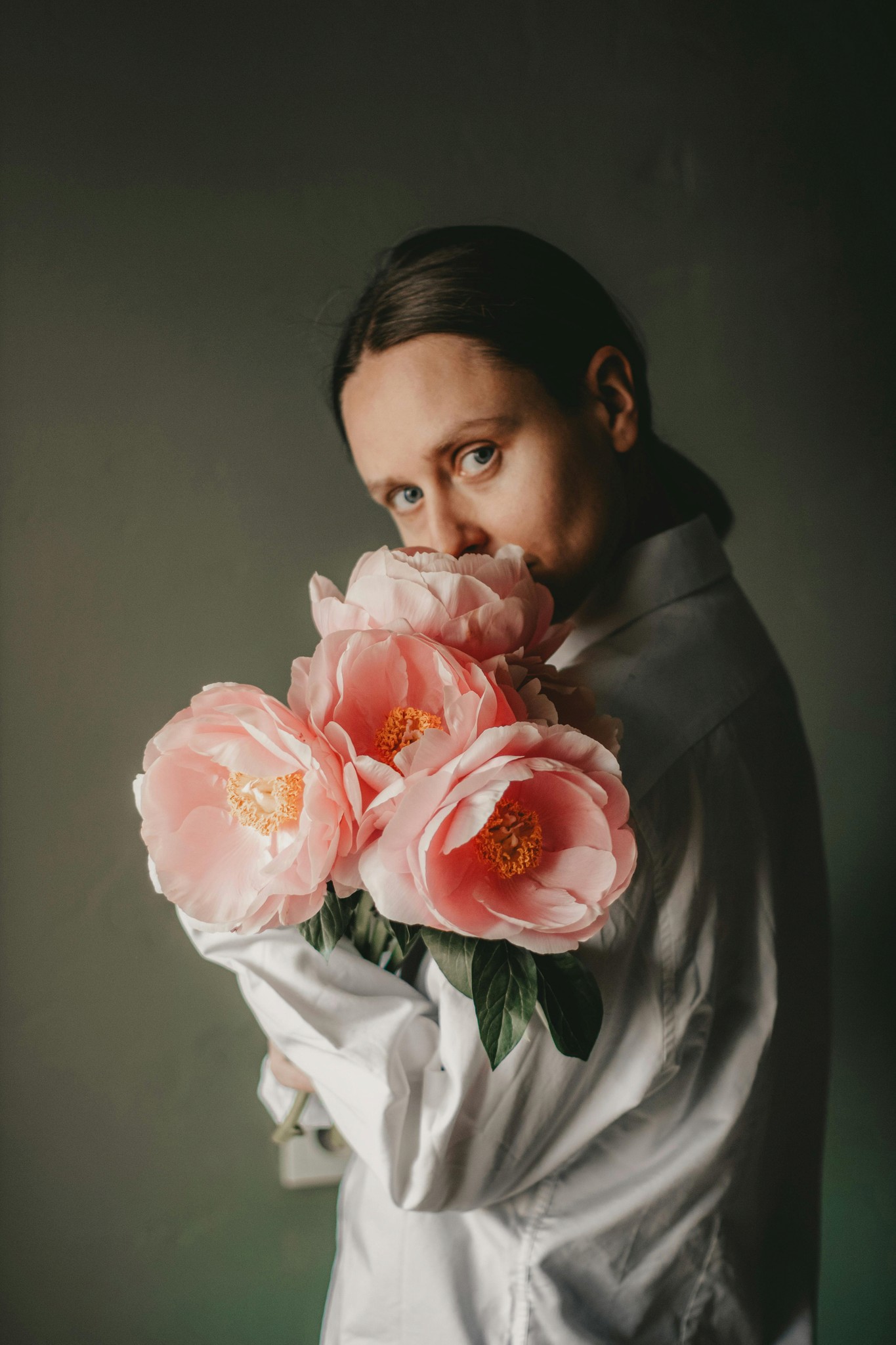 A person in a white shirt peering over a bouquet of large pink peonies against a dark green background.